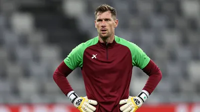 A seasoned goalkeeper in his early 30s, clad in a plain dark red and green training top, stands pitchside with a...