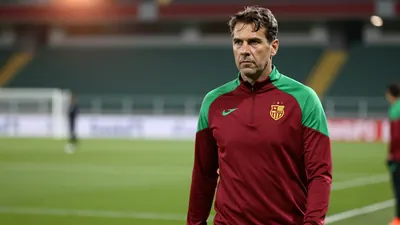 A determined, mid-30s FC Basel assistant coach with a strong build, wearing a plain dark red and green training top...