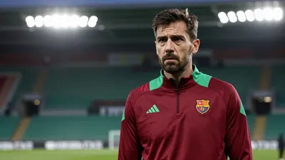A concerned, dark-haired FC Basel staff member in his mid-30s, wearing a plain dark red and green training top, stands...