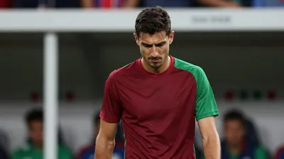 A frustrated, dark-haired FC Basel midfielder, 28, in a plain dark red and green jersey, stands pitchside, eyes cast...