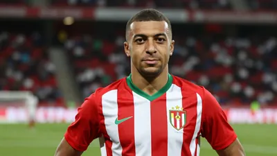 A 24-year-old Moroccan-Belgian winger in a red and white striped jersey, standing confidently on a football pitch with a...