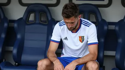 A photorealistic image of an anonymous FC Basel player in their home kit, with a white shirt and blue shorts, sitting on...