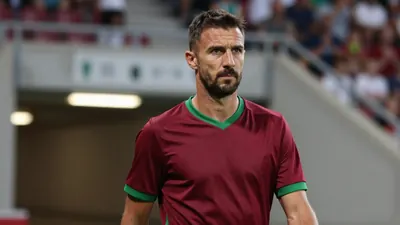 A close-up, waist-up shot of a 35-year-old French coach with a rugged build, wearing a plain, dark red and green...