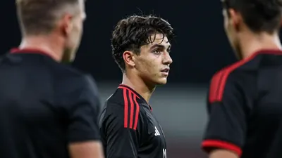 A young defender with dark hair and a narrow face stands with his back to the viewer and looks at the coach, who is...